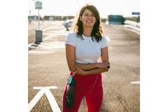 Image A woman wearing a white shirt and pink pants stands in a parking lot, smiling and holding a bottle.
